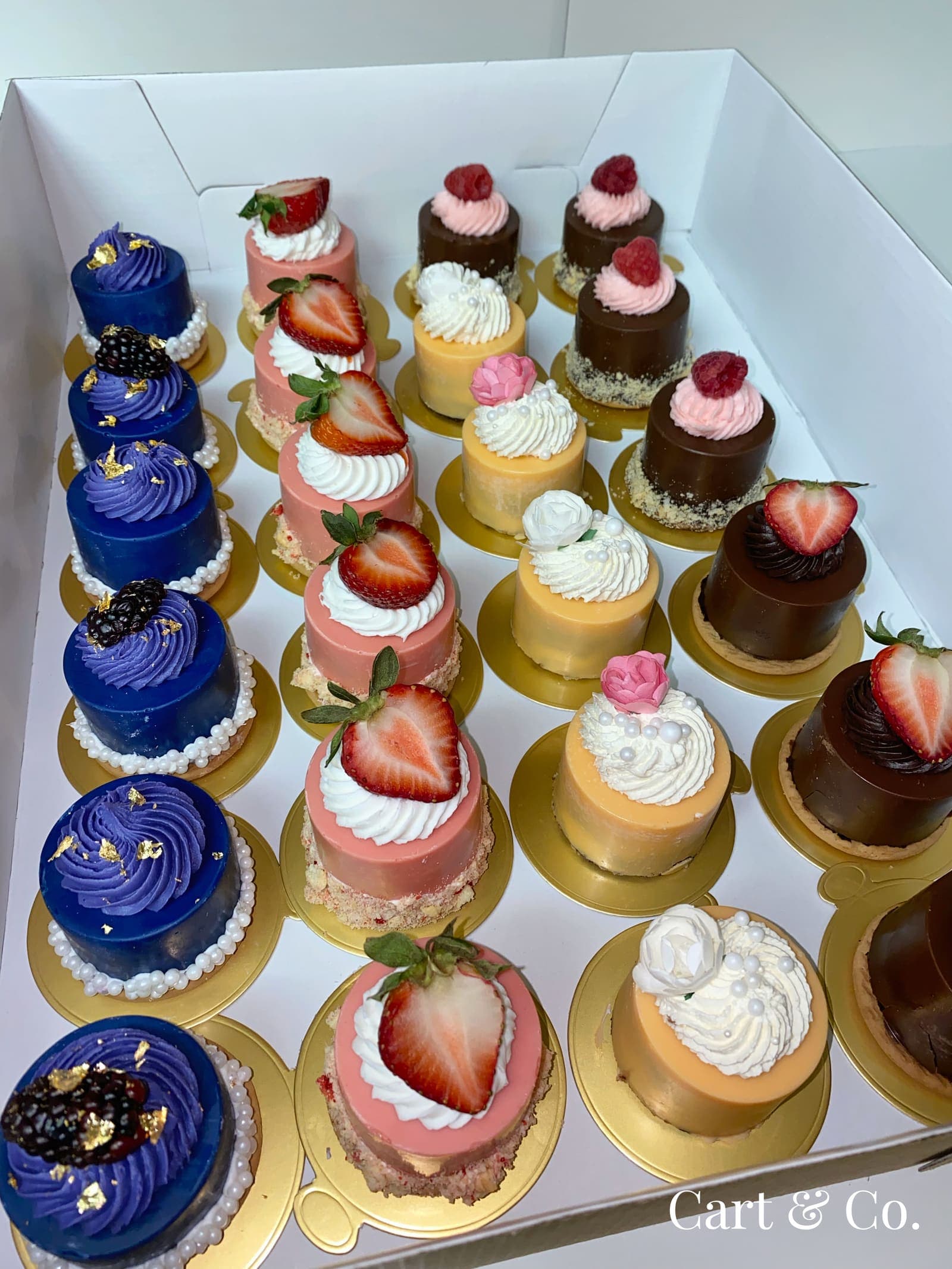 A boxed assortment of blue, pink, gold and chocolate mini cakes topped with fresh fruit, ready for delivery