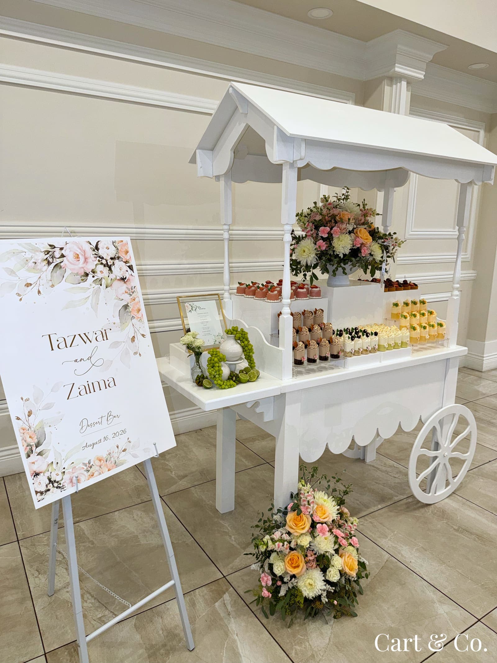 White Cart & Co. dessert cart styled with a floral centerpiece and mini desserts in a ballroom, beside a 'Tazwar and Zaima' welcome sign