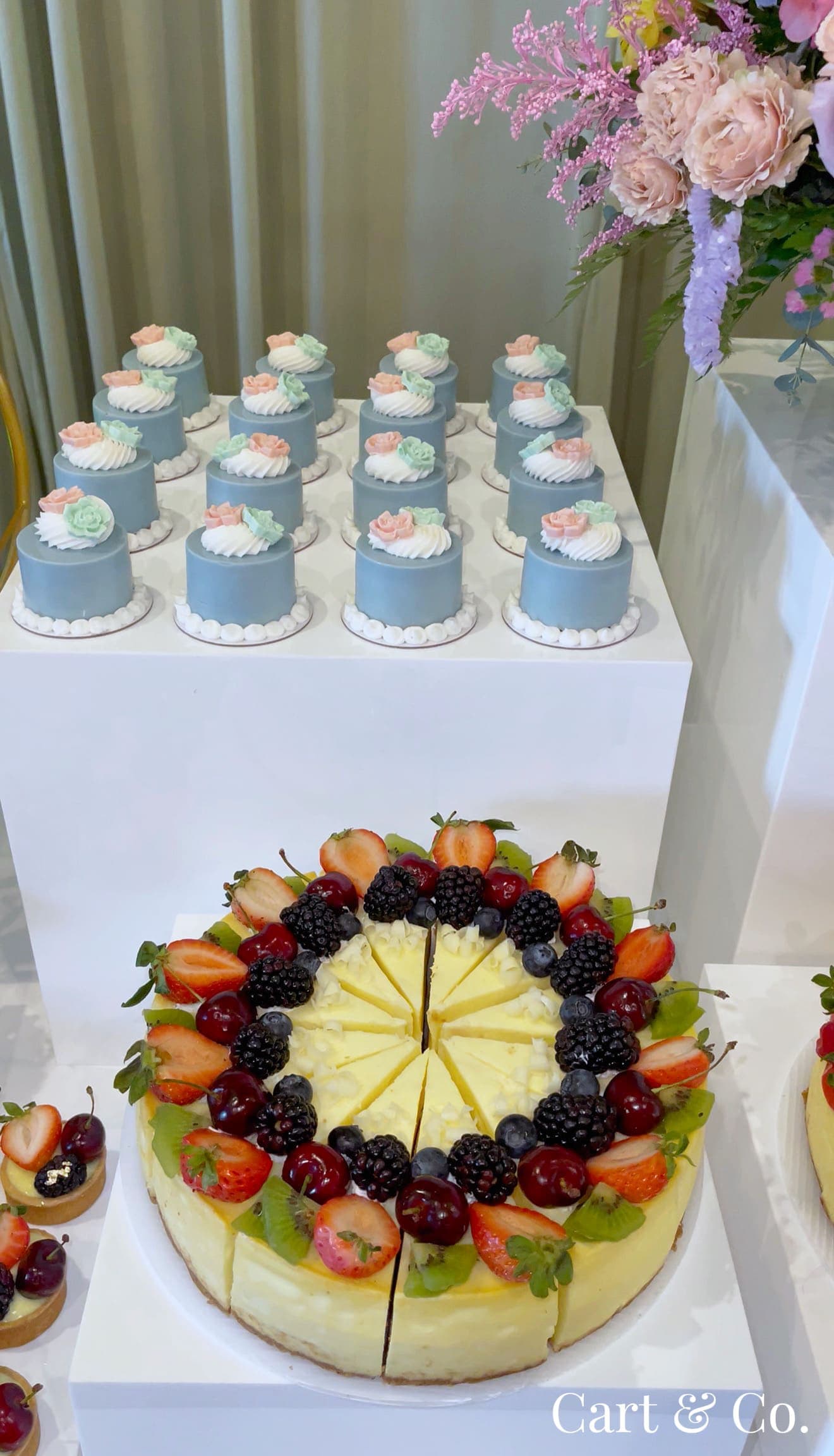 Dusty blue mini cakes with sugar florals beside a fresh berry-topped cheesecake, styled on a dessert table