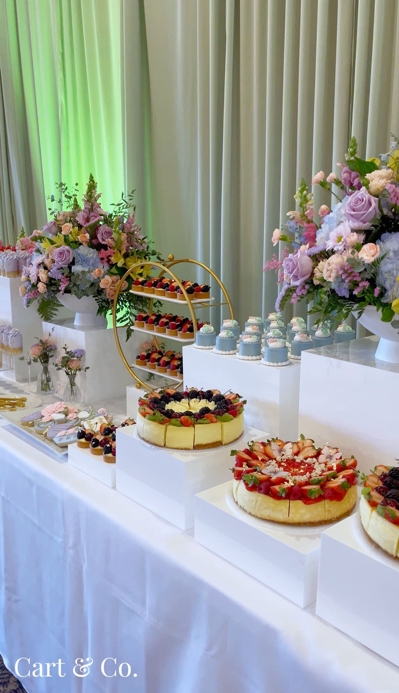 A colorful dessert table with purple, pink and blue mousse cups, berry tarts and a fresh fruit cheesecake