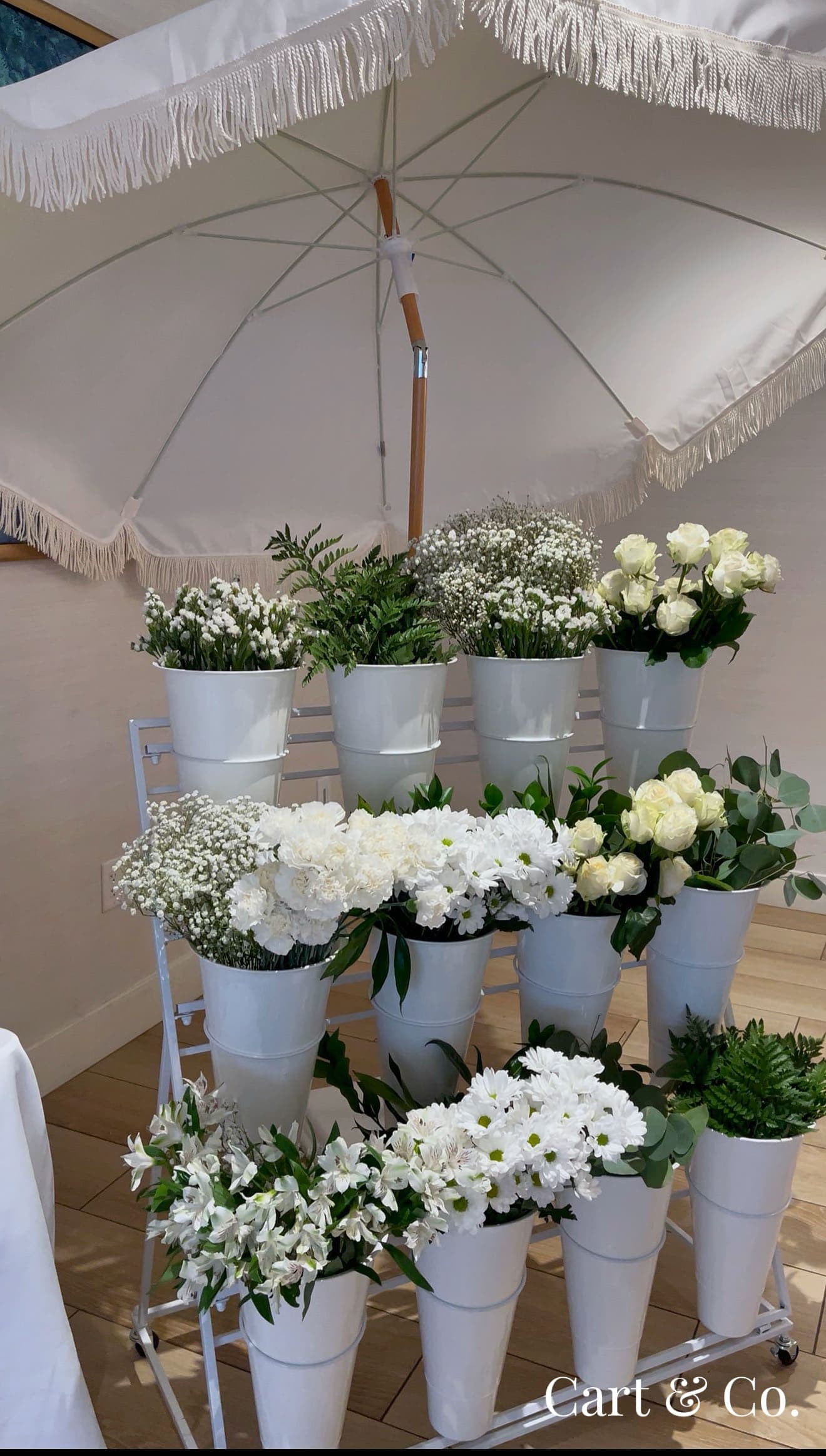 Tiered rack of white roses, daisies and greenery in buckets, staged under a fringed market umbrella before styling