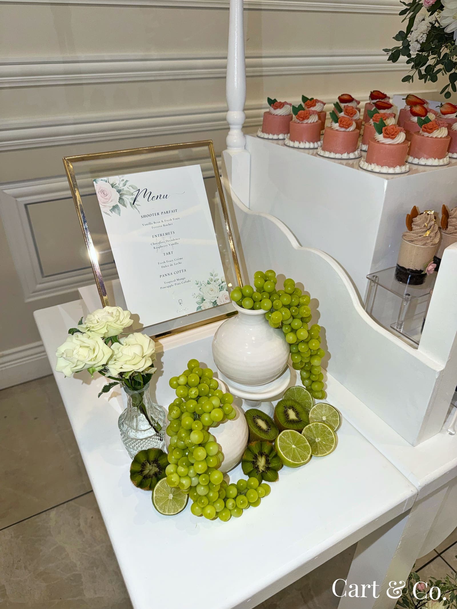 A framed dessert menu beside a garnish of green grapes, kiwi and lime, styled on a Cart & Co. dessert cart