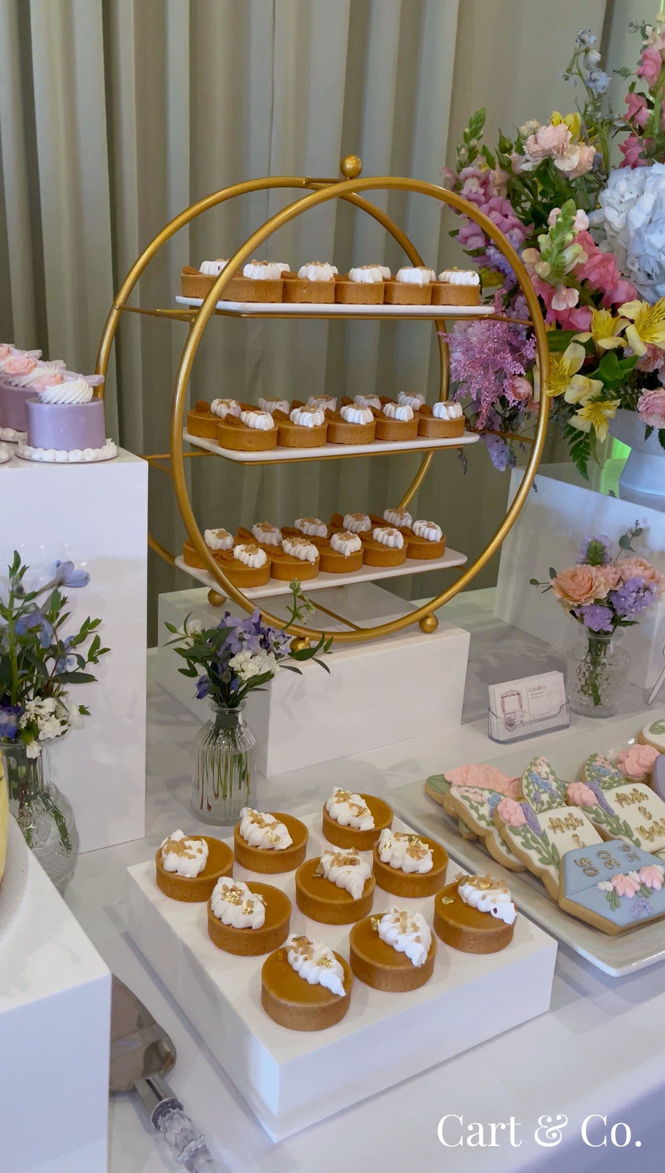 A gold ring-shaped tiered stand with cream-topped tarts, styled beside a pastel floral arrangement