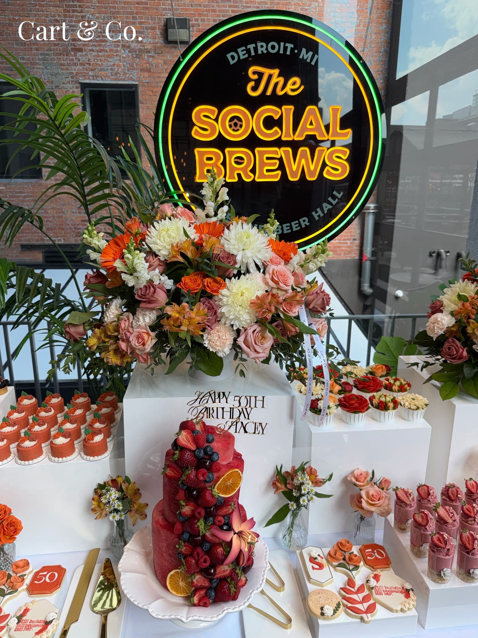 Rooftop 'Happy 50th Birthday' dessert spread with a berry-topped cake, orange rose mini cakes and a floral arrangement beneath 'The Social Brews' neon sign