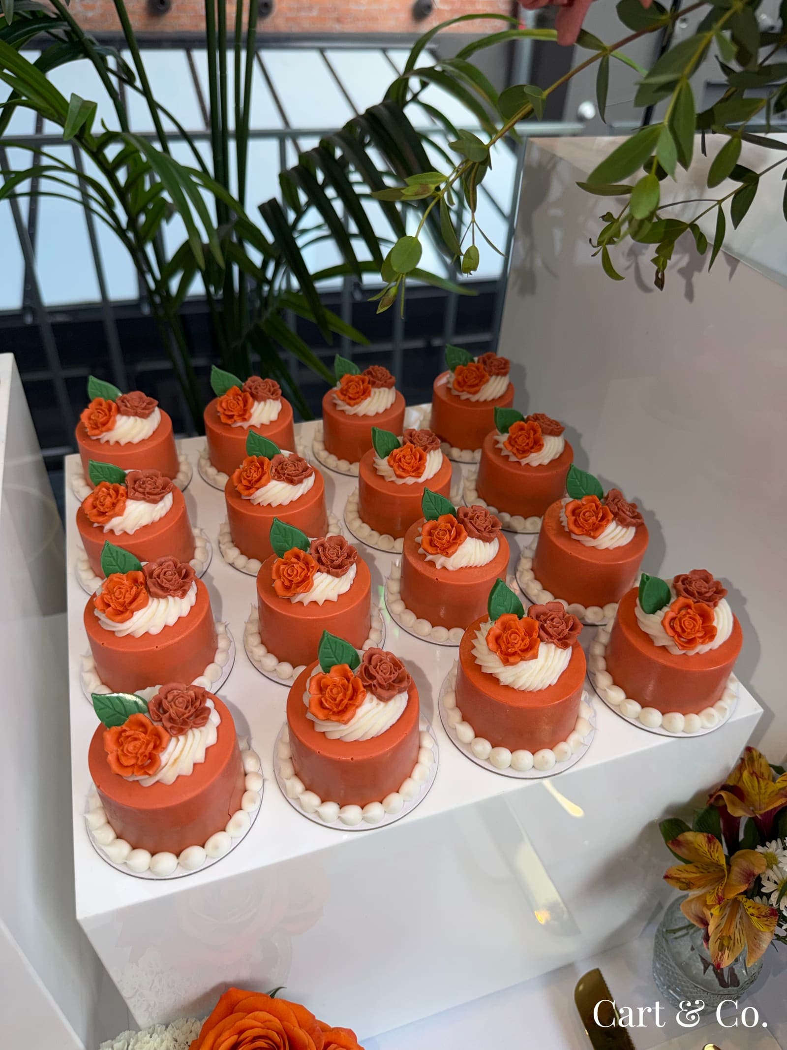 Rows of orange mini cakes topped with sugar roses, styled on a rooftop dessert table against a brick backdrop