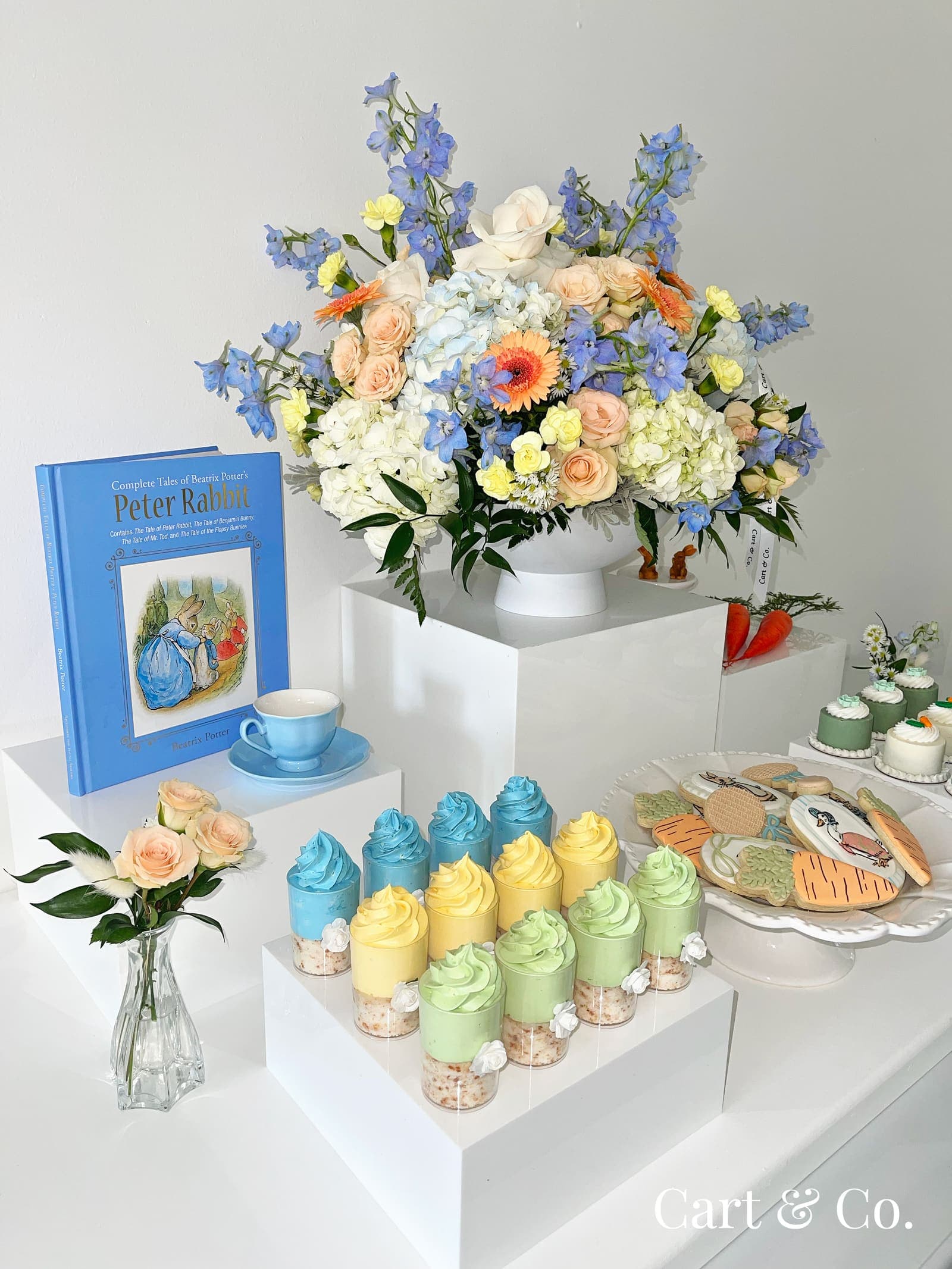 A Beatrix Potter storybook and teacup beside a blue, peach and white floral arrangement on a Peter Rabbit-themed dessert cart