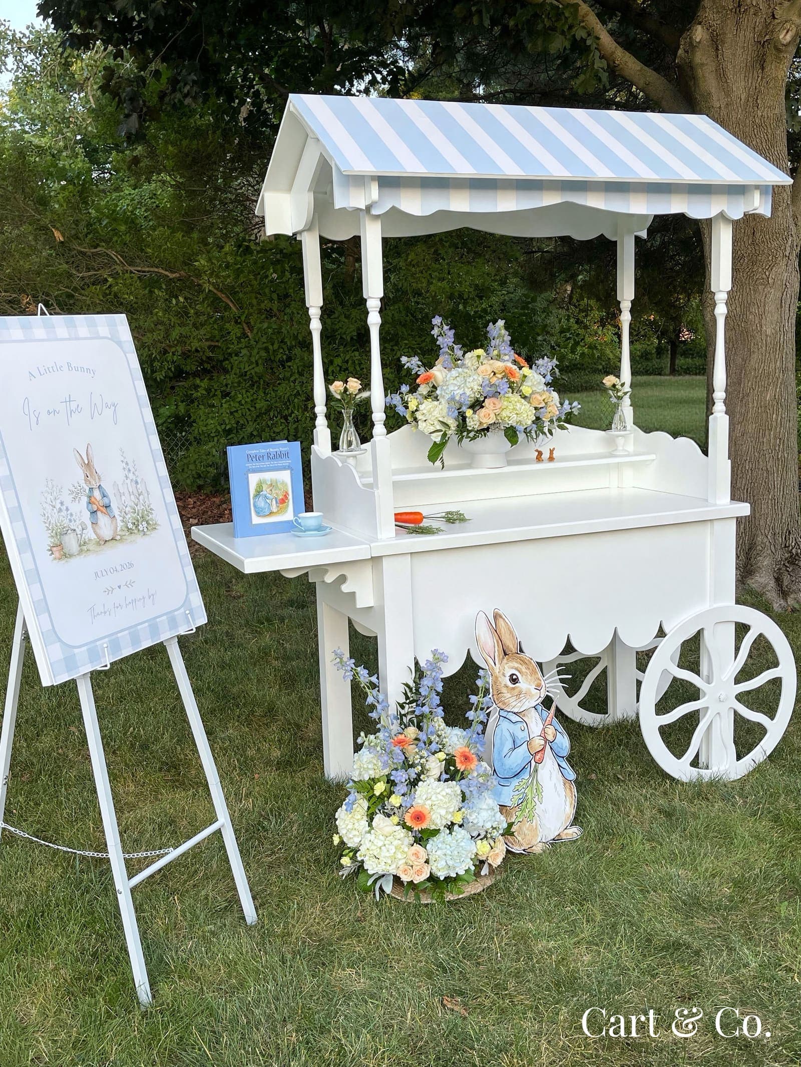A Peter Rabbit-themed dessert cart outdoors with a storybook, teacup and pastel floral arrangement, full scene