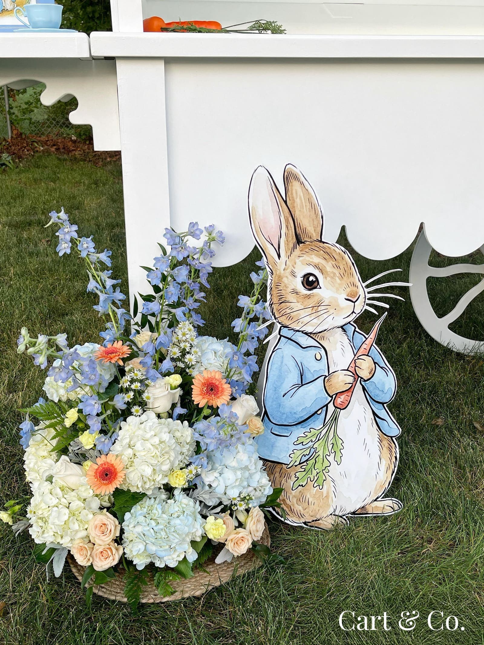 Peter Rabbit cutout holding a carrot beside a blue delphinium and hydrangea arrangement at a baby shower dessert cart