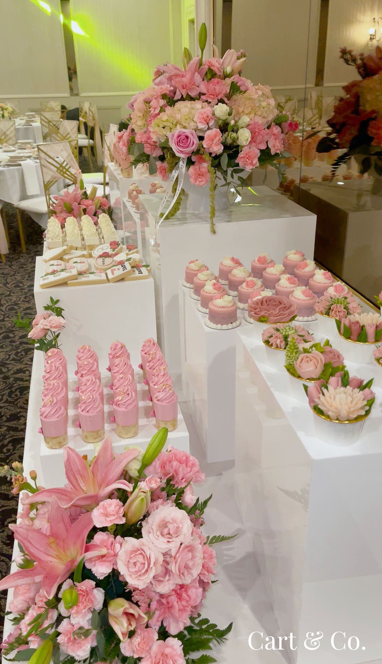 Wide view of a pink bridal shower dessert table in a ballroom, with florals, cookies and mini cakes among gold chiavari chairs