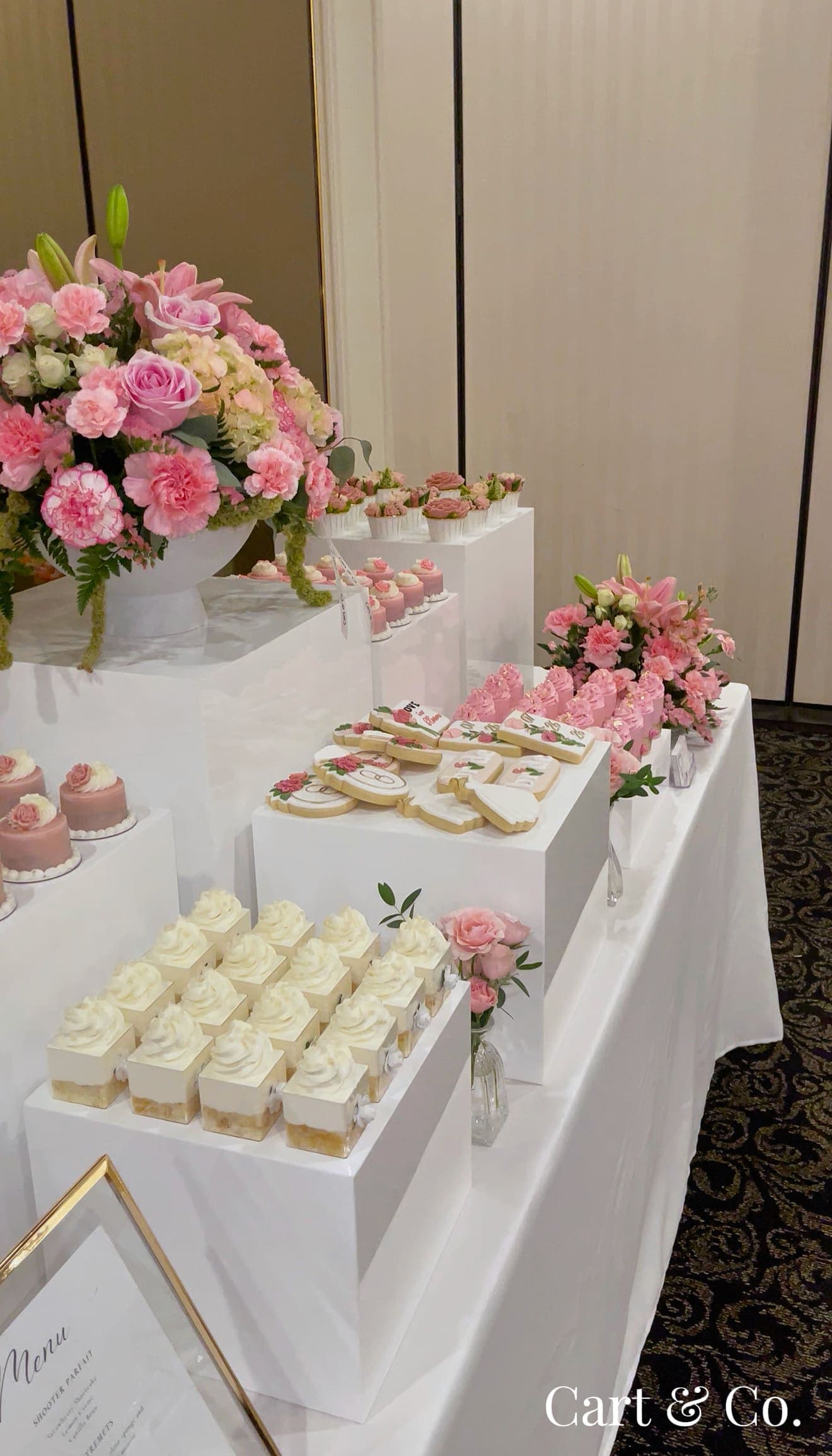 Wide view of a pink ballroom dessert table with a framed menu card, mini cakes, cookies and floral arrangements
