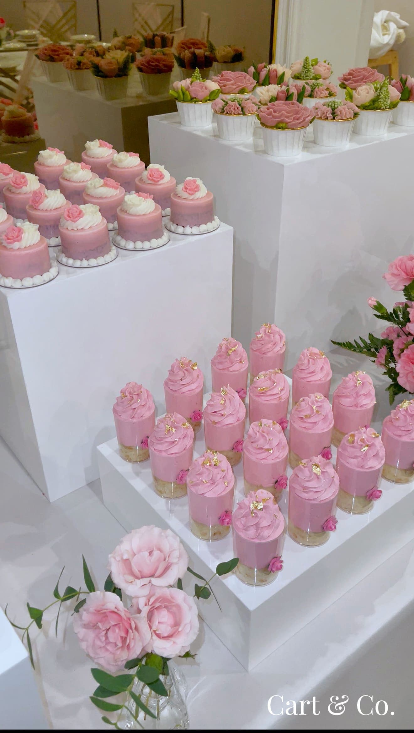 Pink rose-topped cupcakes and mousse cups styled beside a large pink floral arrangement in a mirrored ballroom