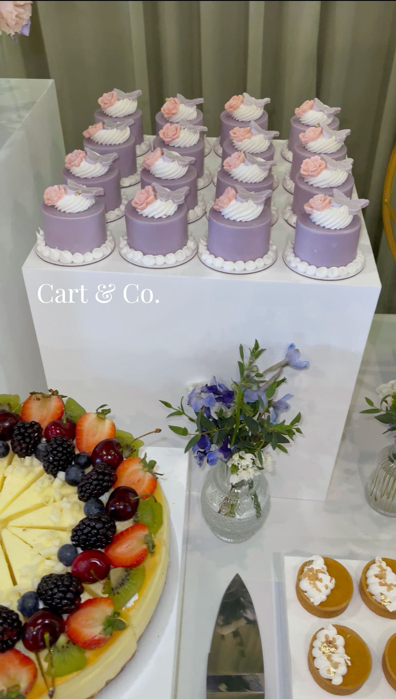 Lavender mini cakes topped with sugar butterflies and a rose, styled beside a fresh fruit cheesecake and blue delphinium for a private event