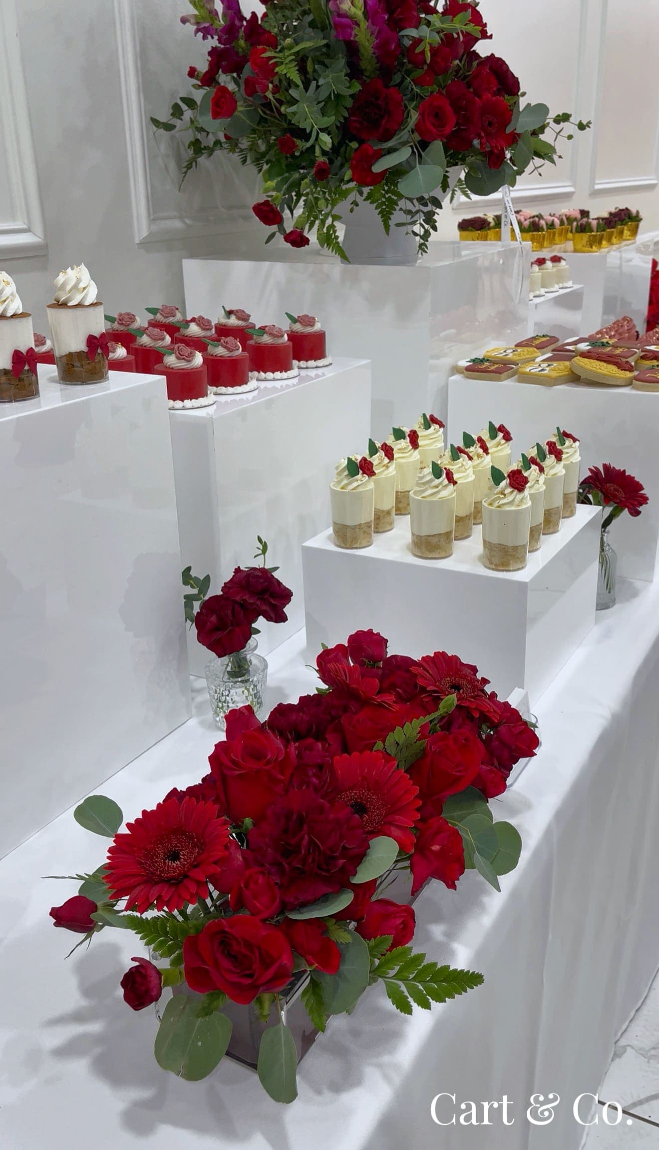 Red rose and gerbera arrangements beside red velvet mini cakes and cookies, styled on a Cart & Co. dessert table