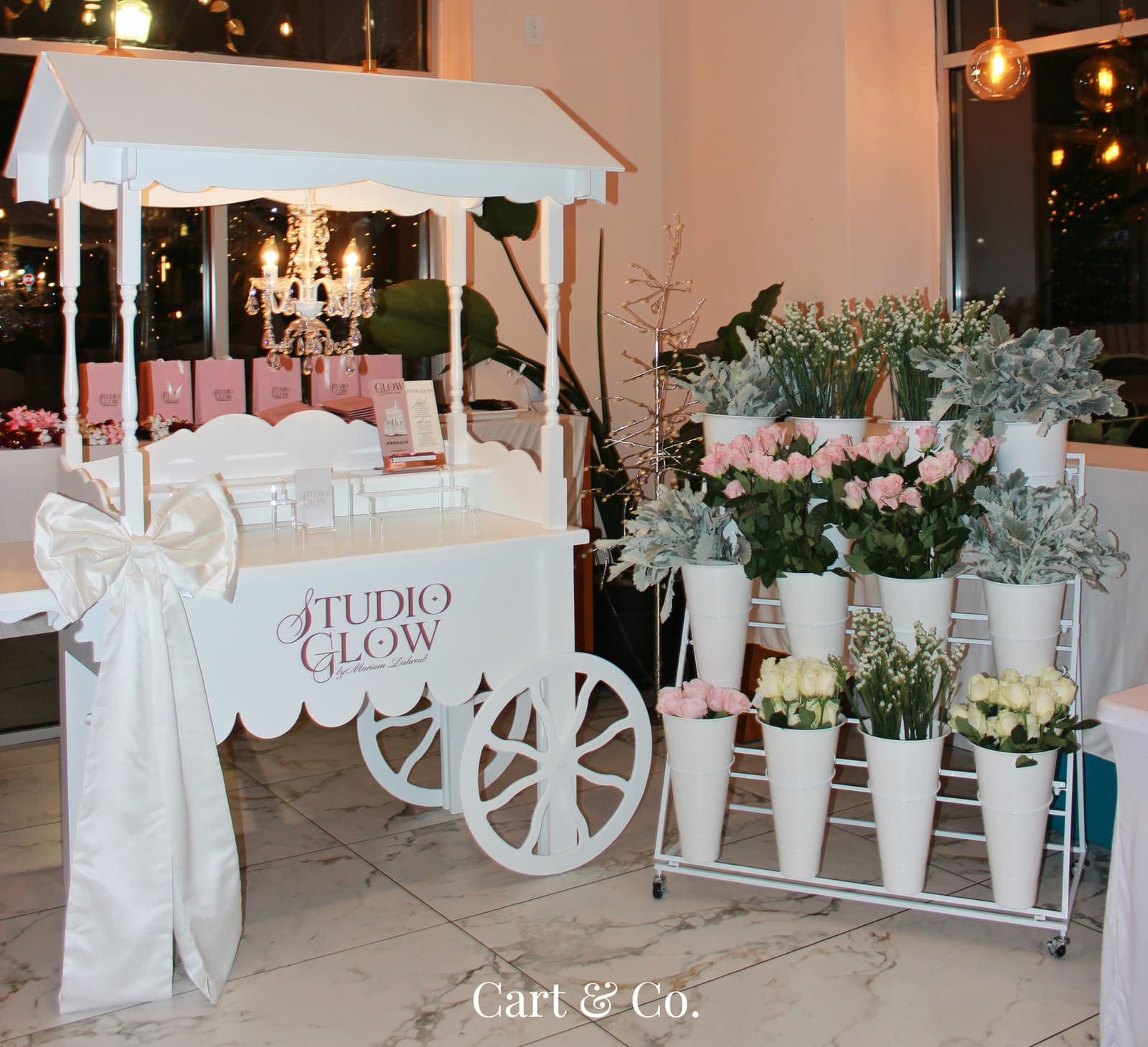 A white Cart & Co. cart custom-branded 'Studio Glow' with a crystal chandelier, ivory satin bow and a tiered flower cart, styled for a product launch
