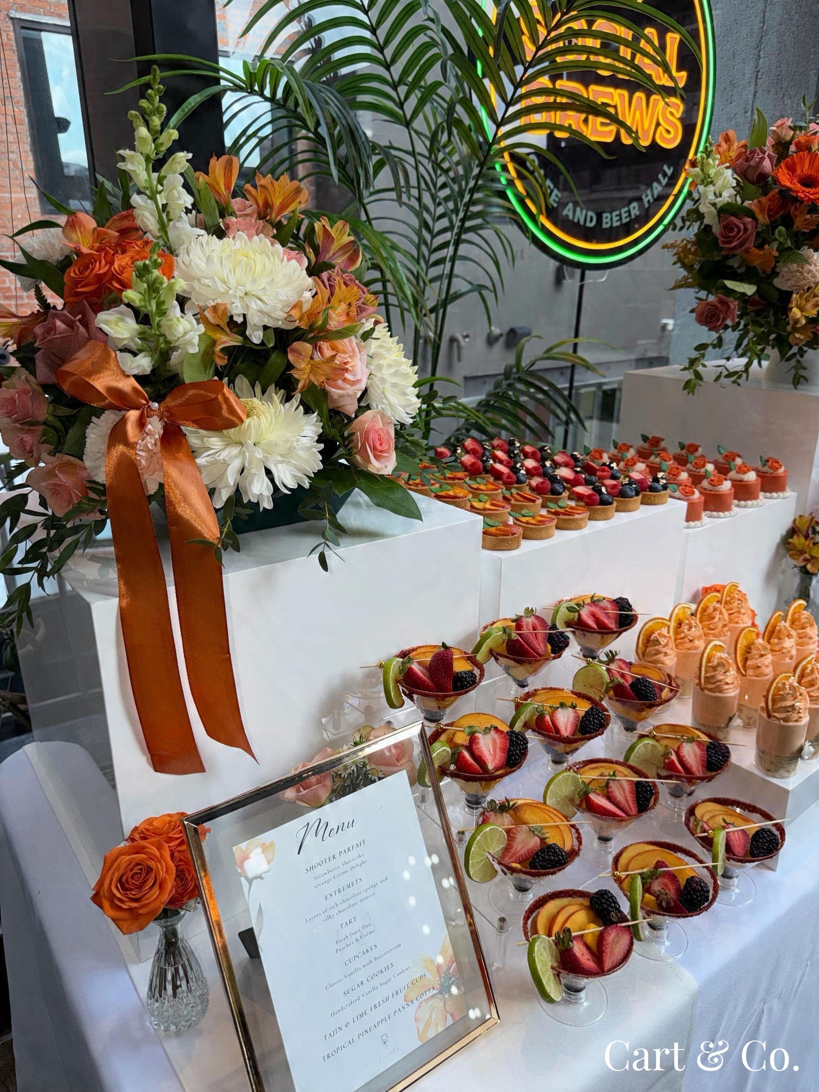 Rooftop 'Happy 50th Birthday' dessert display with berry tarts, orange rose-topped mini cakes, spiced orange mousse cups in margarita glasses and hand-painted birthday cookies, beneath a neon sign
