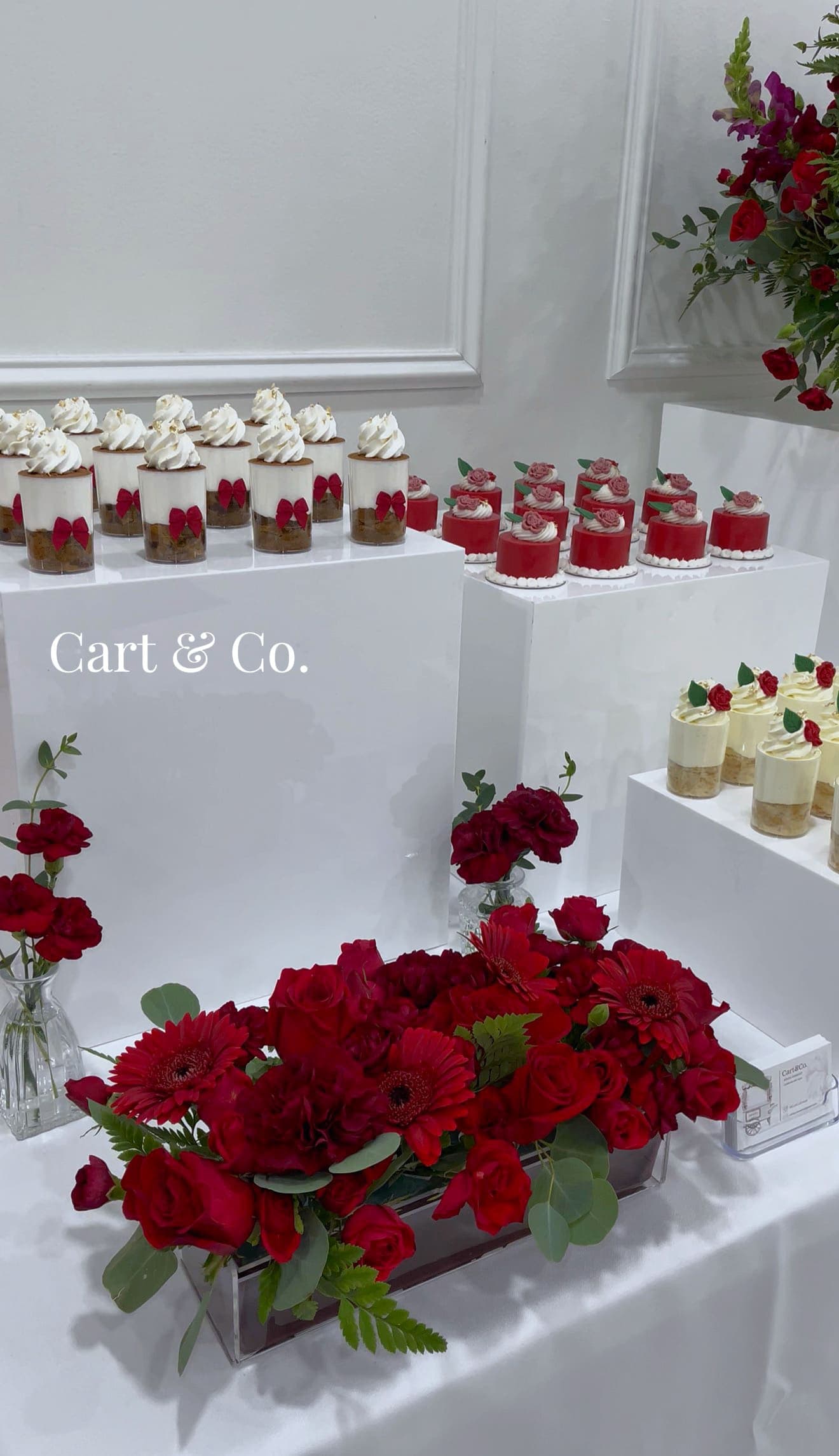 An elegant white Cart & Co. dessert cart with three gold jeweled cake stands and ivory rose swags, styled outdoors under the trees