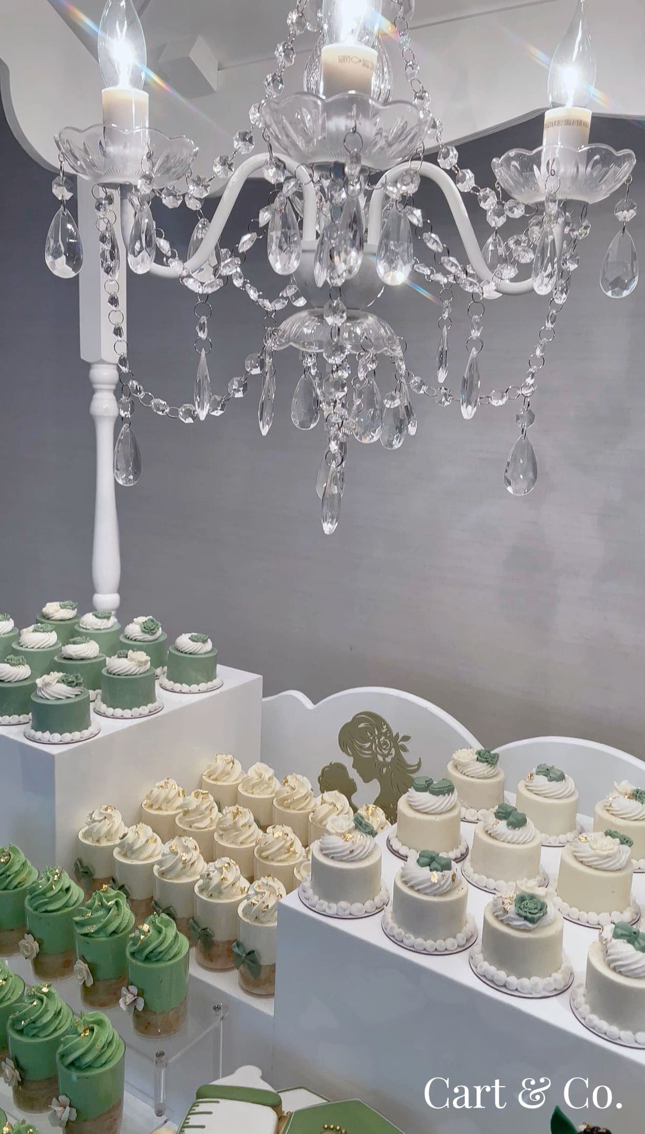 A crystal chandelier over sage green and ivory mini cakes and mousse cups, styled for a baby 'Sweet Reveal'