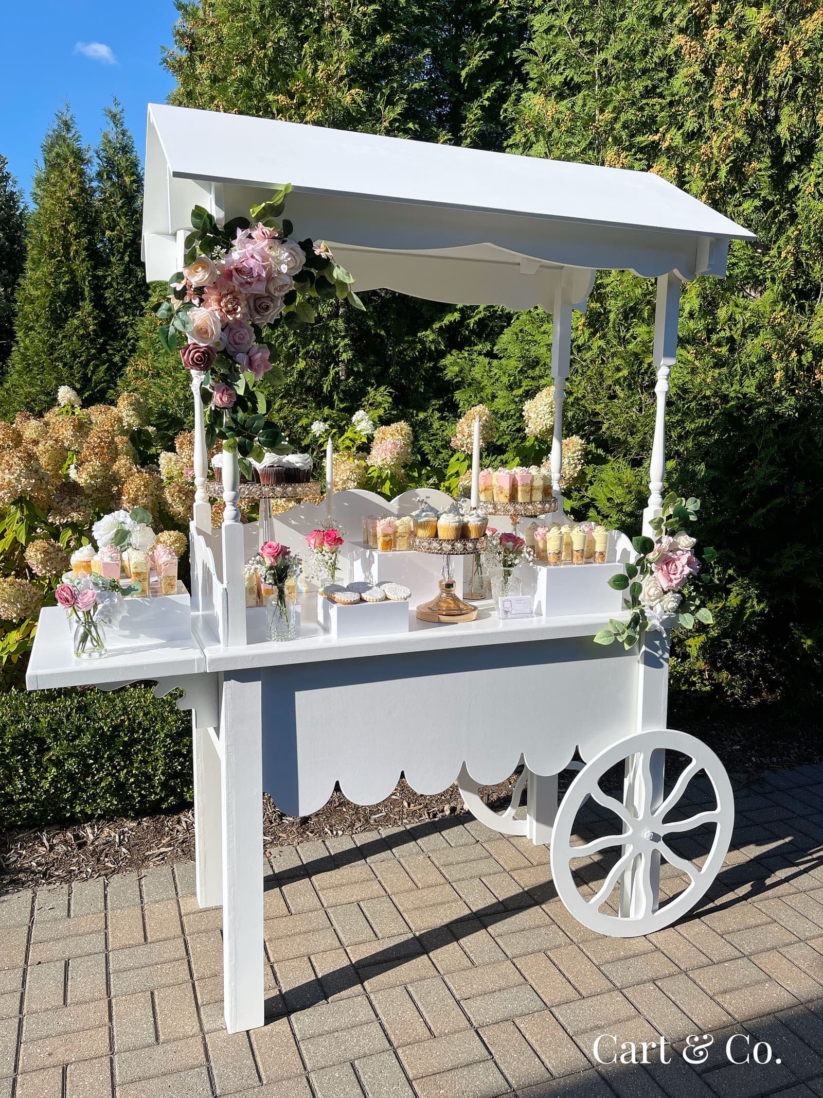 White Cart & Co. dessert cart with pink rose swags, styled among hydrangea bushes with cupcakes and mini desserts