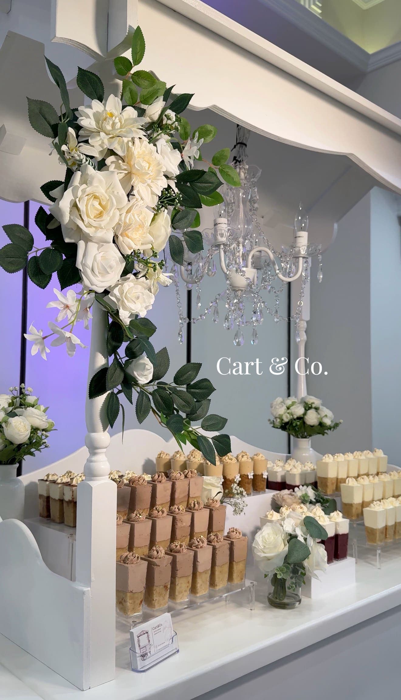 A peach and white floral arrangement beside pink strawberry-topped mini cakes and chocolate mousse cups finished with gold leaf
