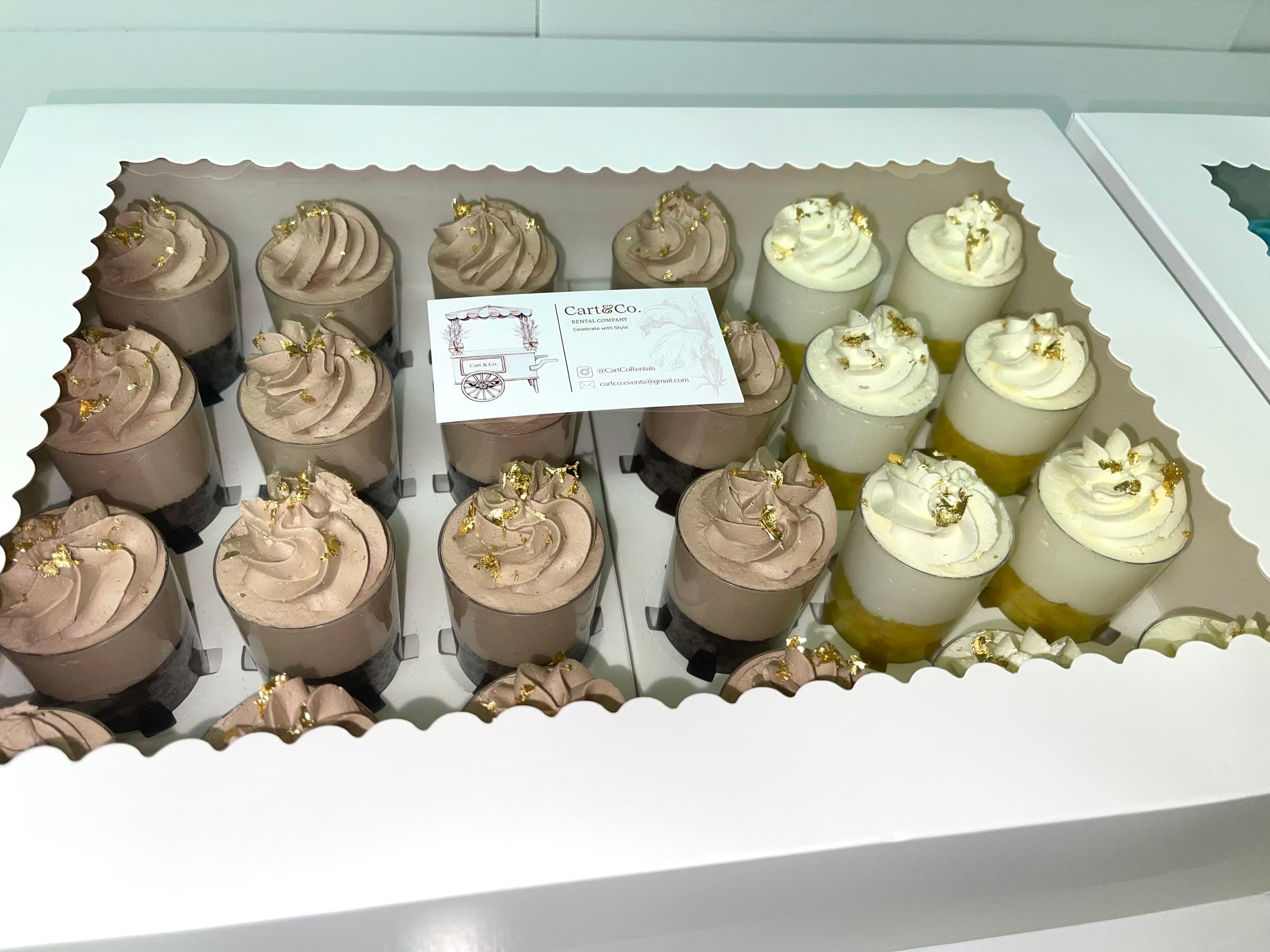 A dozen mousse dessert cups with gold leaf packed in a scalloped white gift box beside a Cart & Co. business card